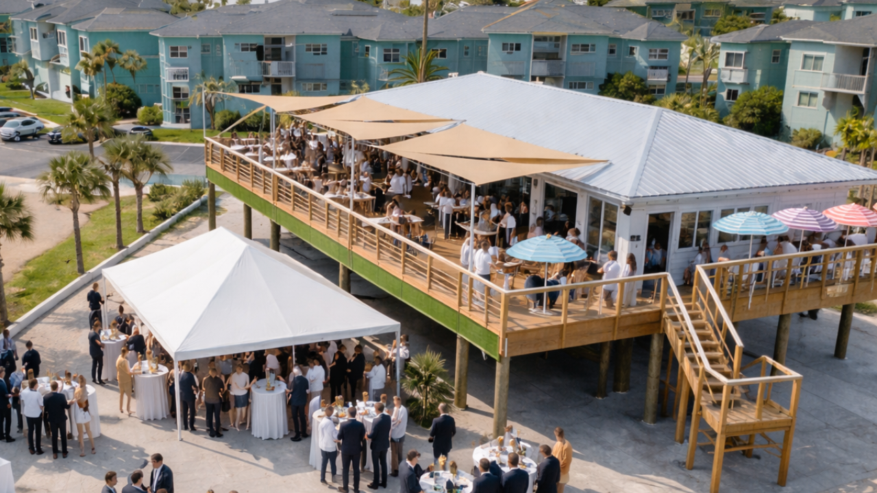 A large outdoor gathering at a beachside venue with guests mingling under tents and umbrellas.
