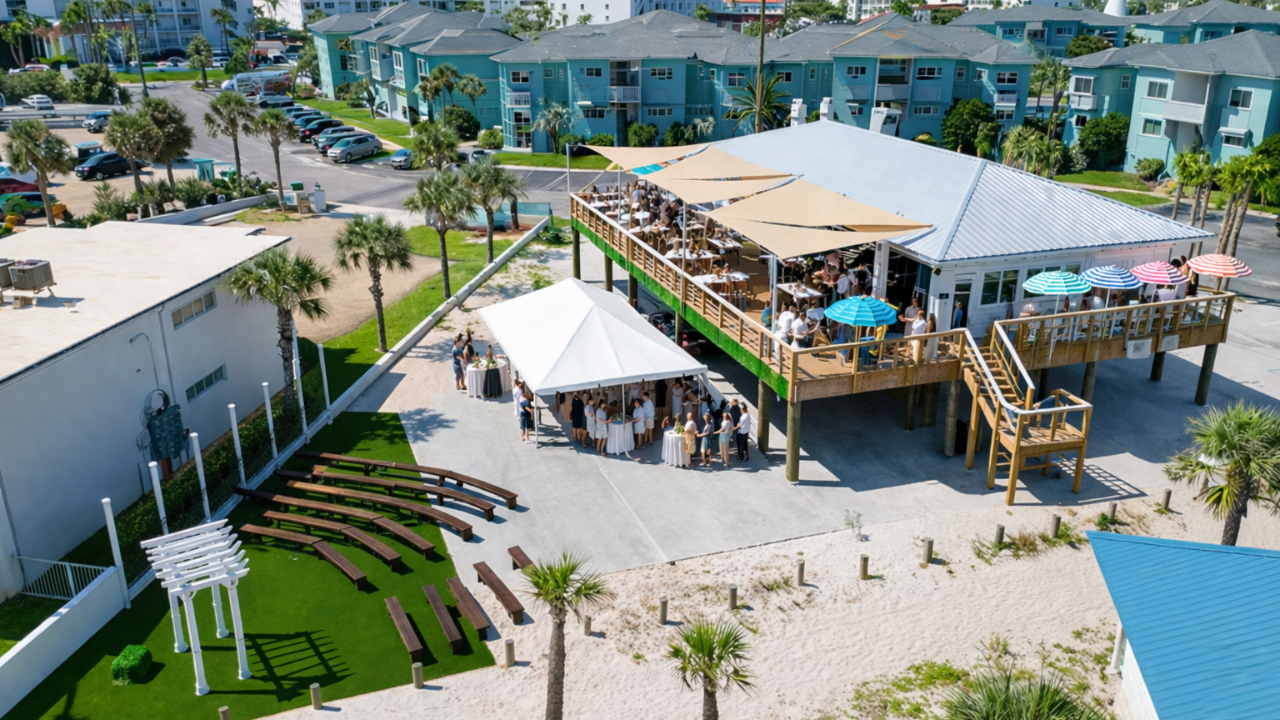 Aerial view of a beachside wedding venue with guests, wooden benches, and a covered deck.