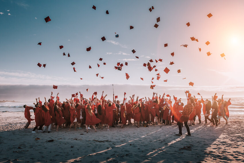 Graduates in red gowns toss caps on a beach at sunset, perfect for a Baby Shower venue.